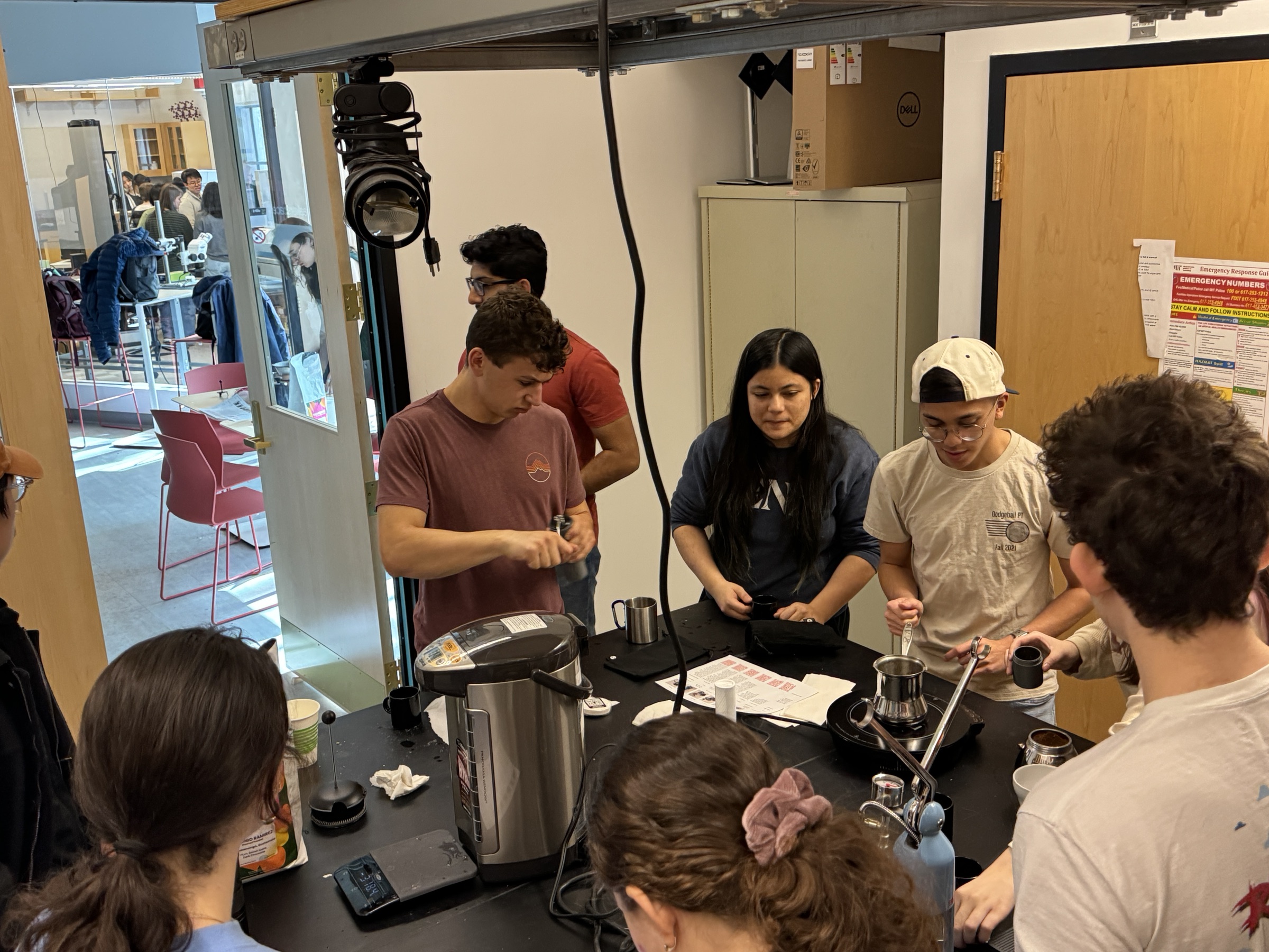 3.000 students gathered around a workbench using kettles, cups, and brewing equipment in food-safe room 8-102B, with the lounge visible through the open door.