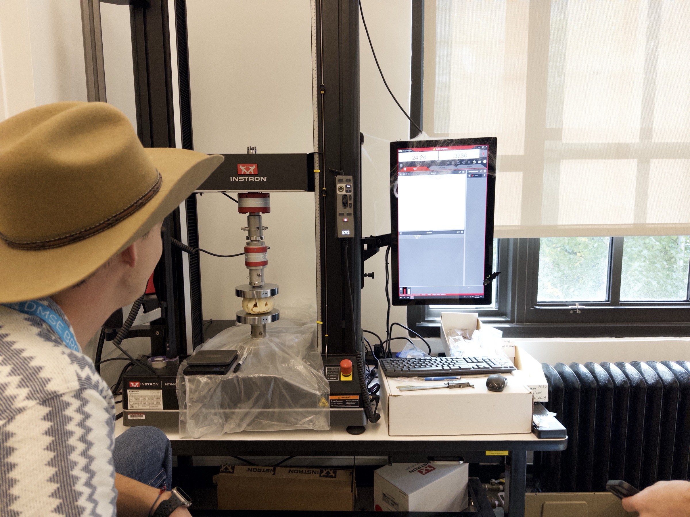 A participant watches a carved pumpkin positioned between circular compression platens in the Breakerspace Instron, with protective sheeting around the test area.